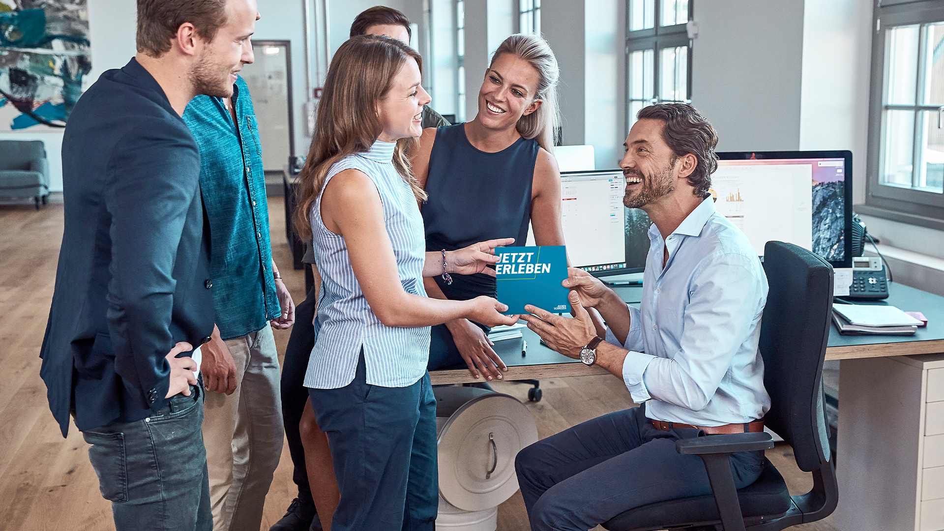 Das Bild zeigt eine Gruppe von fünf Personen, die sich in einem modernen Büro versammelt haben. Drei Männer und zwei Frauen sind sichtbar. Eine Frau steht im Vordergrund und hält ein blaues Rechteck, auf dem „JETZT ERLEBEN“ steht. Sie übergibt es einem Mann, der auf einem Stuhl sitzt und lächelt. Die anderen Personen um sie herum wirken ebenfalls glücklich und lachen. Im Hintergrund sind Schreibtische, Computerbildschirme und große Fenster zu sehen, die den Raum hell und offen wirken lassen. Es scheint eine positive und freundliche Atmosphäre zu herrschen.