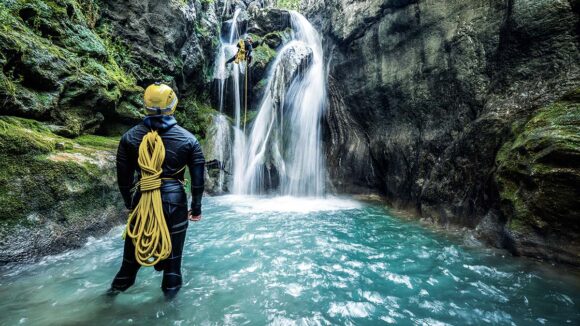 Man steht im Wasser vor einem Wasserfall während sich jemand vom Wasserfall abseilt.